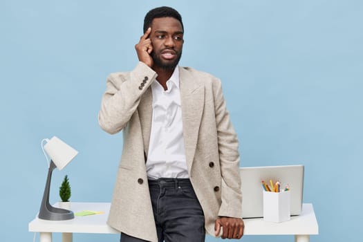 Thoughtful man wearing blazer and white shirt holding headset near ear, standing by desk with laptop, stationery, and lamp, on a plain light blue background. Concept: customer service, communication, professional discussion.