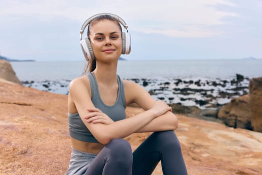 Happy young woman wearing headphones, dressed in a stylish gray sports outfit, relaxing by the beach The serene ocean view creates a calming atmosphere
