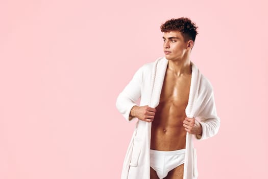 Confident young male model wearing a white bathrobe and underwear, standing against a soft pink backdrop, expressing a casual and relaxed lifestyle concept.
