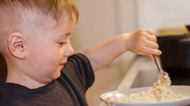 Small child with a spoon eating porridge from a large bowl in the kitchen in the evening. High quality photo