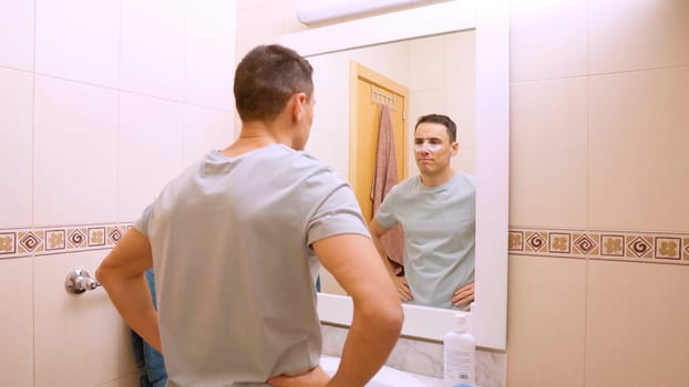 Man taking care of his skin applying anti dark circles patches in front of a bathroom mirror
