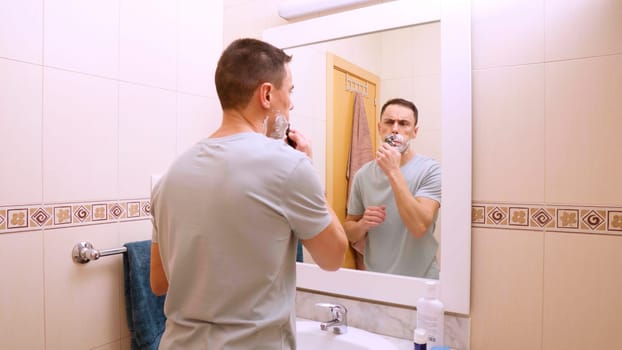Caucasian man shaving his mustache with shaving cream in front of bathroom mirror