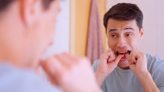 Man using dental floss for cleaning teeth, maintaining oral hygiene in front of bathroom mirror