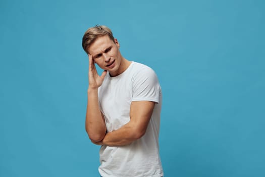 Young man with short hair and casual white t-shirt touching his temple, showing signs of headache or discomfort, standing against bright blue backdrop, facial expression of pain, health and wellness concept, emotional stress, studio shot, isolated on background