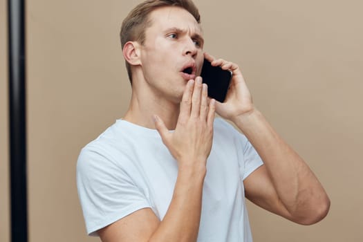 Young man with shocked and worried expression speaking on a mobile phone, dressed casually in a light blue t-shirt, standing against a plain beige background, studio shot. Concept: communication shock
