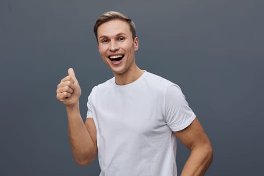 Joyful young man with bright expression wearing white t-shirt, showing thumbs up, standing against a neutral gray studio backdrop. This image captures happiness, confidence, positivity, casual style, and youthful enthusiasm. Concept: optimism and success.