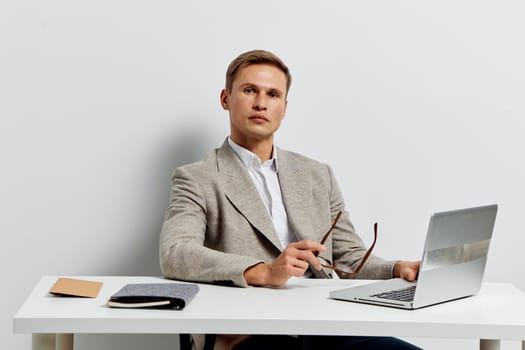 Confident man in business casual outfit sitting at a desk with open laptop and glasses, appearing attentive and engaged, modern indoor workspace with minimalistic decor, neutral hues, professional atmosphere, seated posture, ready for work or discussion, concept of professionalism and productivity.