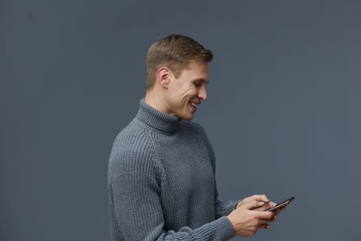 Young smiling man in a cozy gray turtleneck sweater engaging with his mobile phone, standing against a neutral gray background. Captures a moment of connection and modern lifestyle, emphasizing relaxation and technology use. Concept: digital communication.