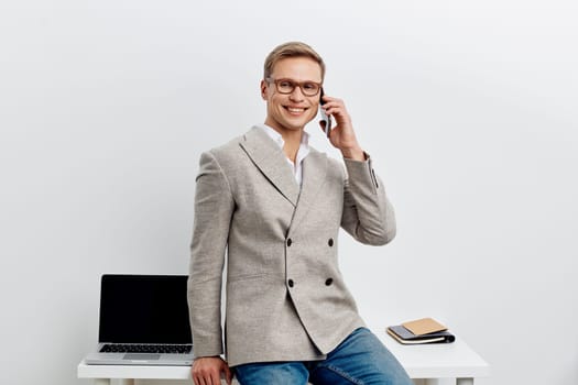 smiling young man talking smartphone wear casual blazer jeans and glasses sit near laptop on desk isolated on white background digital nomad and remote work concept modern technology communication