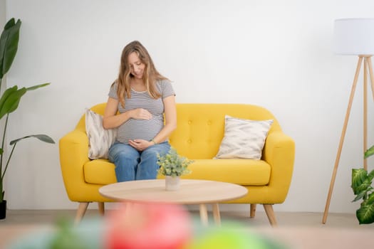 A calm pregnant woman cradles her belly on a cheerful yellow sofa, radiating warmth and tranquility