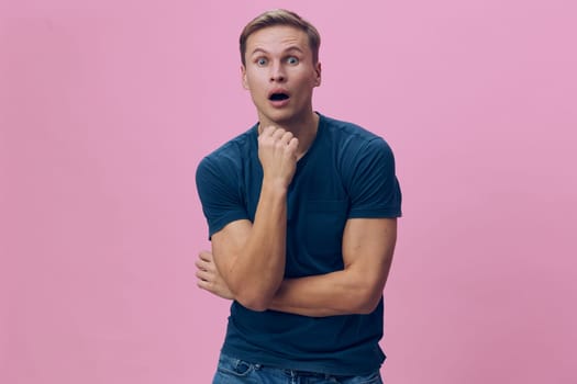 Curious surprised young man with blond hair, wearing blue t-shirt, standing against pink background, looking directly at camera, with hand on chin, expressing amazement, youthful, casual style. Concept: shock, curiosity, emotion, surprise, youth, casual