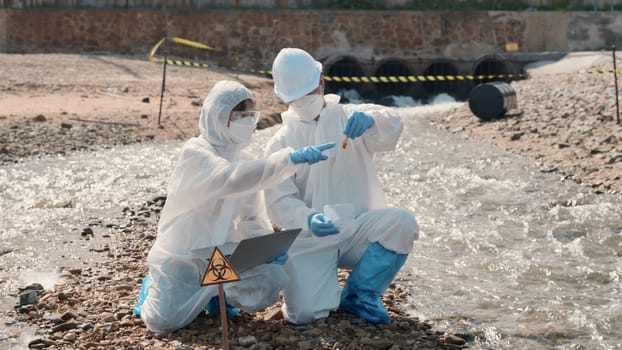 Biologist wear protective suit and mask collects sample of waste water from industry, problem environment, Ecologist sample taken dead fish to inspection and save data to laptop computer, Toxic water