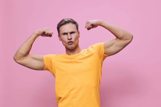 Portrait of a confident young man with a muscular build, dressed in a yellow t-shirt, flexing his biceps to showcase strength and fitness, standing against a pink background. Ideal for health, gym, motivation themes. Concept: body positivity.