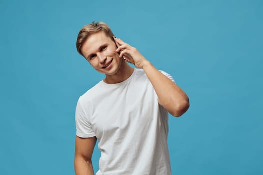 smiling young caucasian man wearing white t-shirt stands against blue background while talking on smartphone showing digital nomad lifestyle and remote work concept modern communication technology