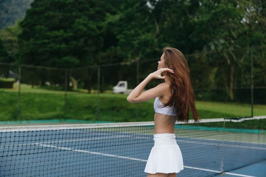Athletic woman in a white tennis outfit stands confidently on a tennis court, surrounded by lush greenery, exuding a vibrant and healthy lifestyle ideal for sports themed content
