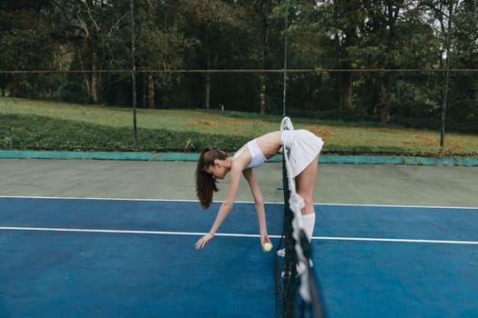 A fit female tennis player in a stylish white outfit, demonstrating concentration and energy while retrieving a tennis ball, with a lush green background highlighting an active lifestyle