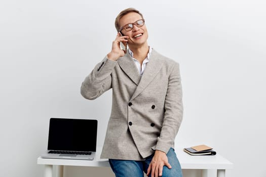 smiling man talking phone wear casual gray blazer and glasses sit on white table with laptop and notebook isolated on white background digital nomad concept and work in metaverse communication technology and business lifestyle