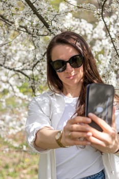 Beautiful mid aged woman takes self portrait with her phone in the blooming trees with flowers in the garden