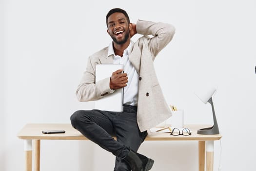Happy man sitting on desk in contemporary office environment holding tablet, cheerful expression, relaxed posture, casual business style, minimalistic workspace, positive work atmosphere