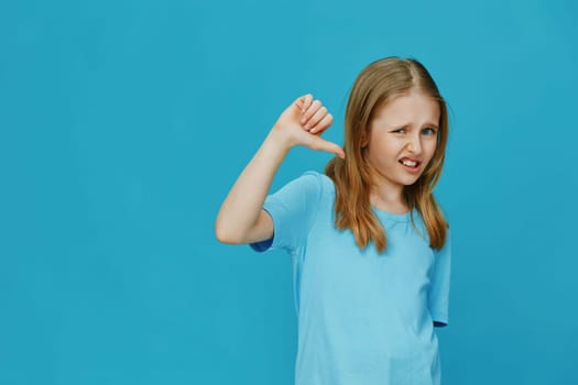 A young girl expresses her disappointment with a thumbs down gesture on a vibrant blue background She wears a light blue shirt, capturing a mixture of emotions and playful energy
