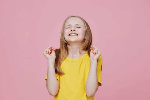 Joyful young girl in a yellow t shirt with crossed fingers, smiling brightly against a pink background This image conveys excitement, happiness, and a sense of celebration
