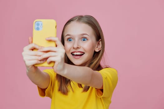 Excited girl capturing a selfie with her smartphone, wearing a bright yellow t shirt, against a playful pink background, radiating joy and youthful spirit