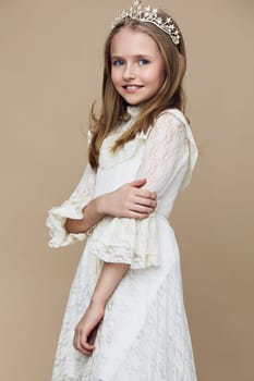 Smiling young girl wearing a beautiful white dress with lace details and a crown, posed gracefully on a soft beige background, symbolizing elegance, joy, and childhood innocence