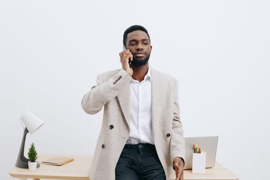 Confident man with dark skin dressed in smart casual clothing holding a mobile phone in hand, standing in a contemporary office space with minimal decor. Concept: professional communication and modern work environment.