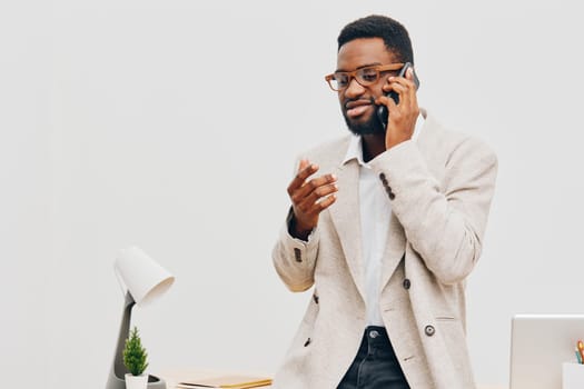 Professional man in beige blazer and white shirt smiling confidently while talking on mobile phone in office setting with clean decor, white wall, and desk accessories. Business lifestyle, communication, modern workspace.