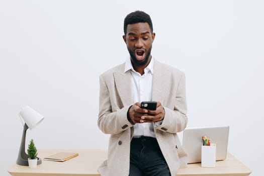 Confident young man of African descent in smart casual clothing, standing at a desk with a laptop, surprised and engaged, holding a smartphone, minimalist office environment, neutral tones, indoor setting. Concept: modern work life