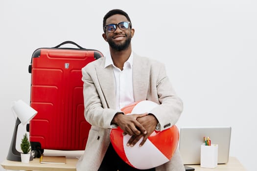 A cheerful man with dark skin, stylish glasses, and a beige blazer holding a paper heart in a contemporary office space with a red suitcase, laptop, and office supplies, conveying themes of travel, love, and positivity. Concept: celebration of relationships and adventures
