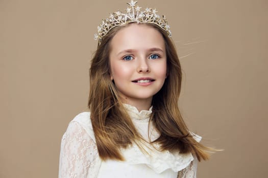A joyful young girl wearing a delicate white lace dress and a sparkling crown poses beautifully against a soft beige background, exuding innocence and grace