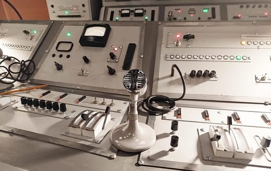 A vintage broadcasting studio showcases a control panel filled with various buttons, knobs, and levers. A classic microphone sits prominently at the center, symbolizing communication.