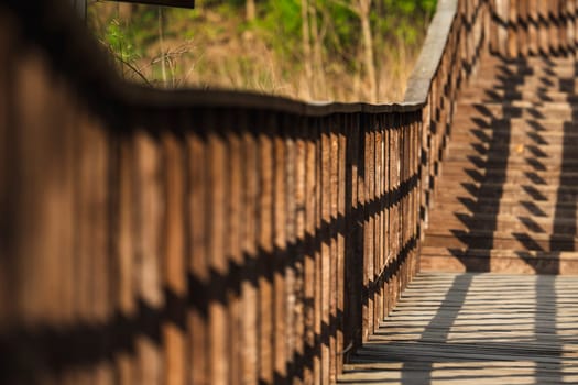 Close-up view of a wooden railing and stairs with diagonal shadow patterns in warm sunlight, surrounded by dry grass and green forest