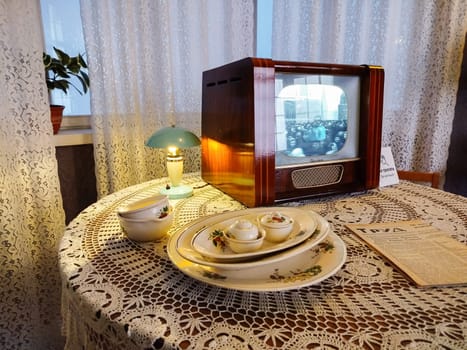 A cozy vintage living room setup features a wooden television, floral dishware on a lace tablecloth, a green lamp, and a newspaper. It evokes nostalgia and timeless charm.