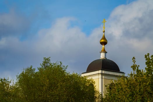A golden onion-shaped dome with an Orthodox cross rises above lush green trees under a partly cloudy blue sky in spring.