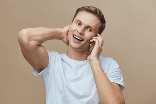 Cheerful young man with short hair dressed in a white casual t-shirt, enjoying a pleasant phone conversation and smiling brightly. He is holding his neck in a relaxed manner, with a neutral beige background, conveying positive emotions and communication. Concept: everyday life, happiness.