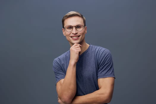 Happy young man with glasses dressed casually in a blue t-shirt, smiling and looking at the camera on a solid gray background. Concept: confidence and positivity.