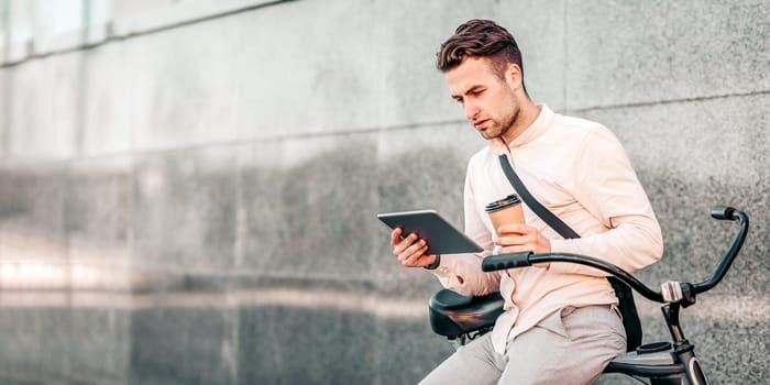A young man sits casually on his bike, engaged with a tablet and holding a coffee cup. He appears focused and relaxed in a modern urban environment during the day, copy space