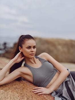 Athletic female model in gray clothing embodies a fit lifestyle, exuding calm emotions while relaxing on a rock near the sea in a serene coastal setting