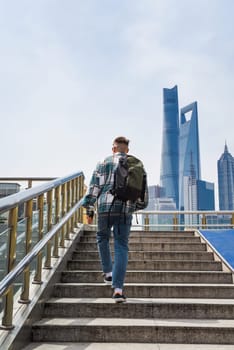 A young man wearing a plaid shirt, jeans, and sneakers walks up stairs with a backpack in Shanghai's Pudong, modern skyscrapers towering behind him