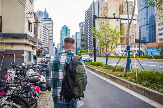 Russian male tourist with backpack explores modern Shanghai alone, walking past scooter, car, and high-rise building on city street