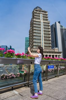 Young woman takes a photo on a pedestrian bridge in Pudong, Shanghai, surrounded by flowers and modern skyscrapers on a sunny spring day