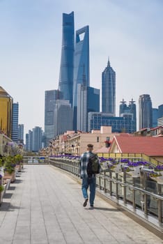 A male tourist walks on a flower-filled bridge in Pudong, Shanghai, with skyscrapers like Shanghai Tower and Shanghai World Financial Center (SWFC) behind him.