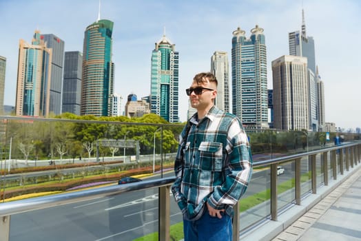 A Russian male traveler in glasses and shirt stands on a Pudong bridge, Shanghai, with modern skyscrapers in the background. Sunny urban scene with copy space.