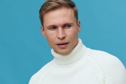 Young caucasian man wearing white turtleneck sweater looking thoughtful aside with serious expression isolated on blue background. Mental health and gadgets concept with calm studio portrait and clean style.