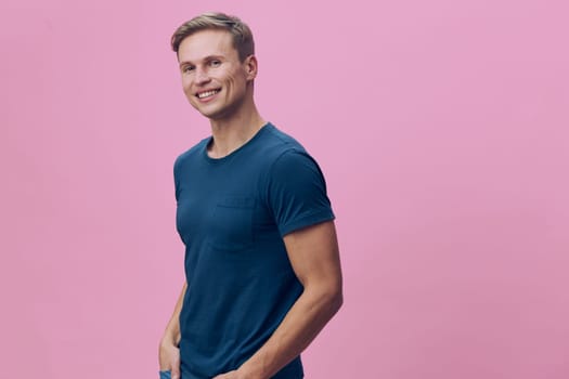 Happy young man in blue casual t-shirt standing against pink background, smiling confidently, relaxed pose, bright studio setting. Suitable for lifestyle, positivity, health themes.