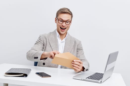 Happy cheerful man sitting at a desk with laptop, surprised while opening an envelope in bright office environment with minimal decor. Concept: workplace positivity and success