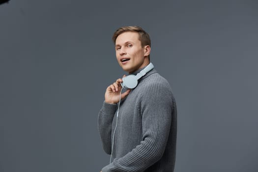 Smiling man enjoying music with over-ear headphones, dressed in a cozy gray sweater, standing against a solid gray background. The image captures relaxation, modern lifestyle, and personal entertainment. Concept: music enjoyment