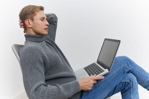 Young man relaxing in a modern indoor setting, dressed in warm casual clothing, leaning back with a laptop resting on his lap, peaceful expression, minimalist background, emphasizing comfort and digital lifestyle.